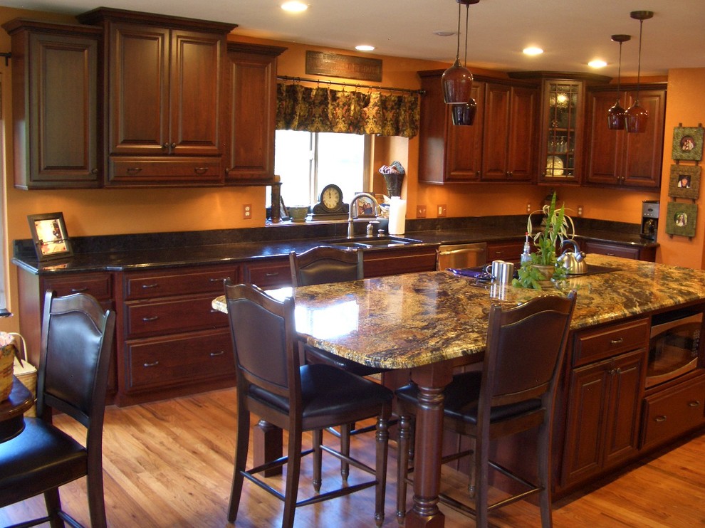 This is an example of a large traditional u-shaped eat-in kitchen in Denver with raised-panel cabinets, dark wood cabinets, with island, an undermount sink, granite benchtops, stainless steel appliances, light hardwood floors, beige floor, black splashback, stone slab splashback and black benchtop.