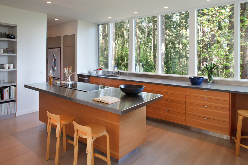 This is an example of a mid-sized contemporary galley kitchen in Seattle with stainless steel benchtops, flat-panel cabinets, medium wood cabinets, stainless steel appliances, a single-bowl sink, light hardwood floors and with island.