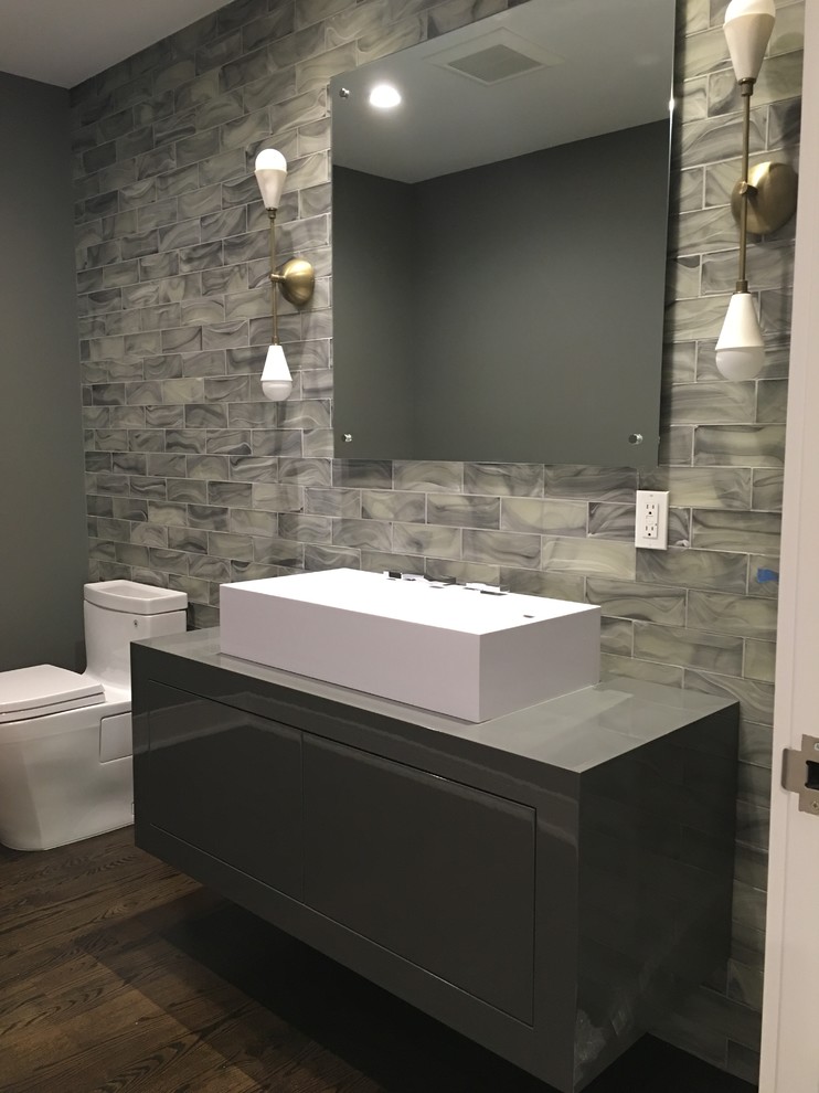 This is an example of a large modern powder room in New York with flat-panel cabinets, grey cabinets, a one-piece toilet, gray tile, glass tile, grey walls, dark hardwood floors, a vessel sink, wood benchtops and brown floor.