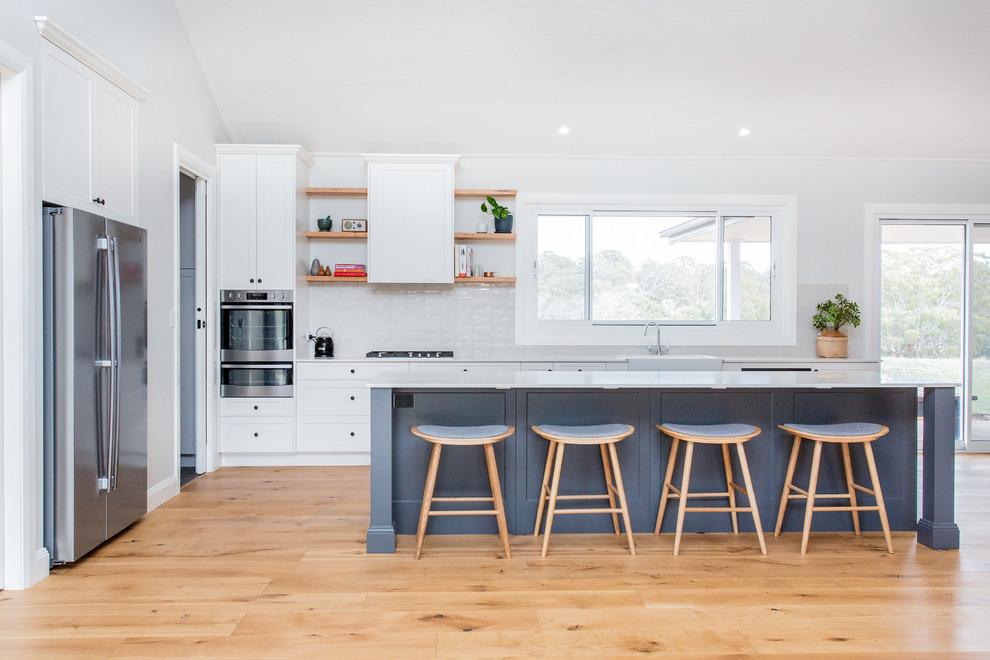 Mid-sized transitional galley open plan kitchen in Other with a farmhouse sink, shaker cabinets, white cabinets, quartz benchtops, white splashback, subway tile splashback, stainless steel appliances, light hardwood floors, with island, brown floor and white benchtop.