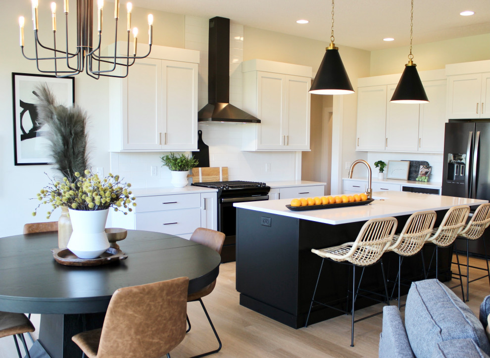 This is an example of a large transitional l-shaped open plan kitchen in Other with an undermount sink, flat-panel cabinets, black cabinets, quartz benchtops, white splashback, subway tile splashback, black appliances, light hardwood floors, with island and white benchtop.