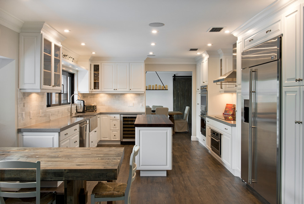Transitional u-shaped eat-in kitchen photo in Miami with a farmhouse sink, raised-panel cabinets, white cabinets, wood countertops, white backsplash and stainless steel appliances