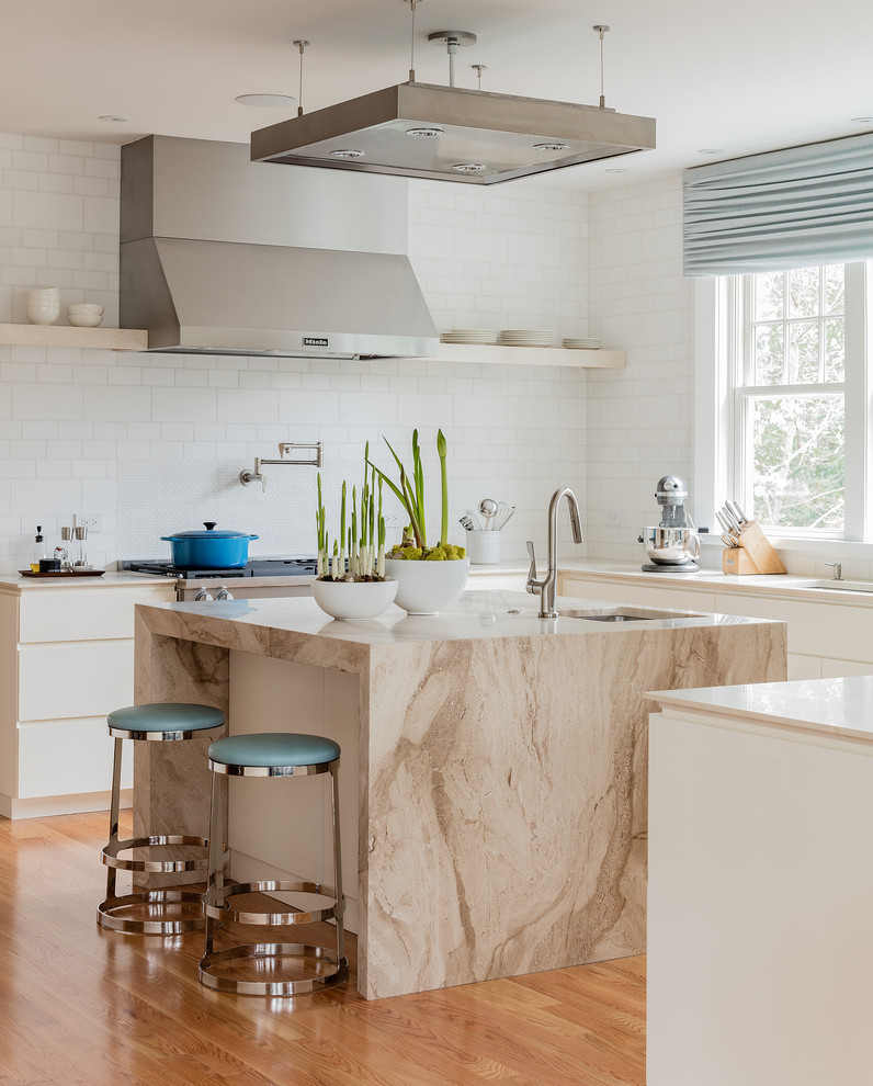 Photo of a beach style u-shaped kitchen in Boston with an undermount sink, flat-panel cabinets, white cabinets, marble benchtops, white splashback, stainless steel appliances, medium hardwood floors, with island and brown floor.