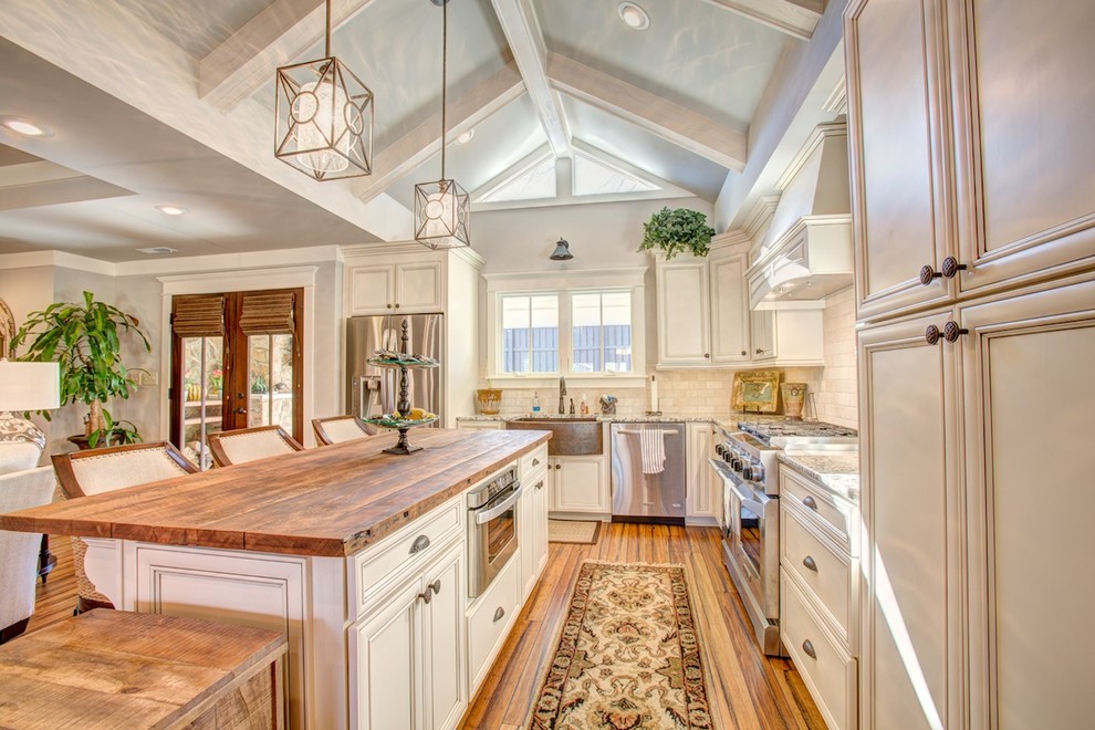 Inspiration for a mid-sized timeless l-shaped bamboo floor open concept kitchen remodel in Birmingham with a farmhouse sink, flat-panel cabinets, white cabinets, wood countertops, beige backsplash, stainless steel appliances and travertine backsplash