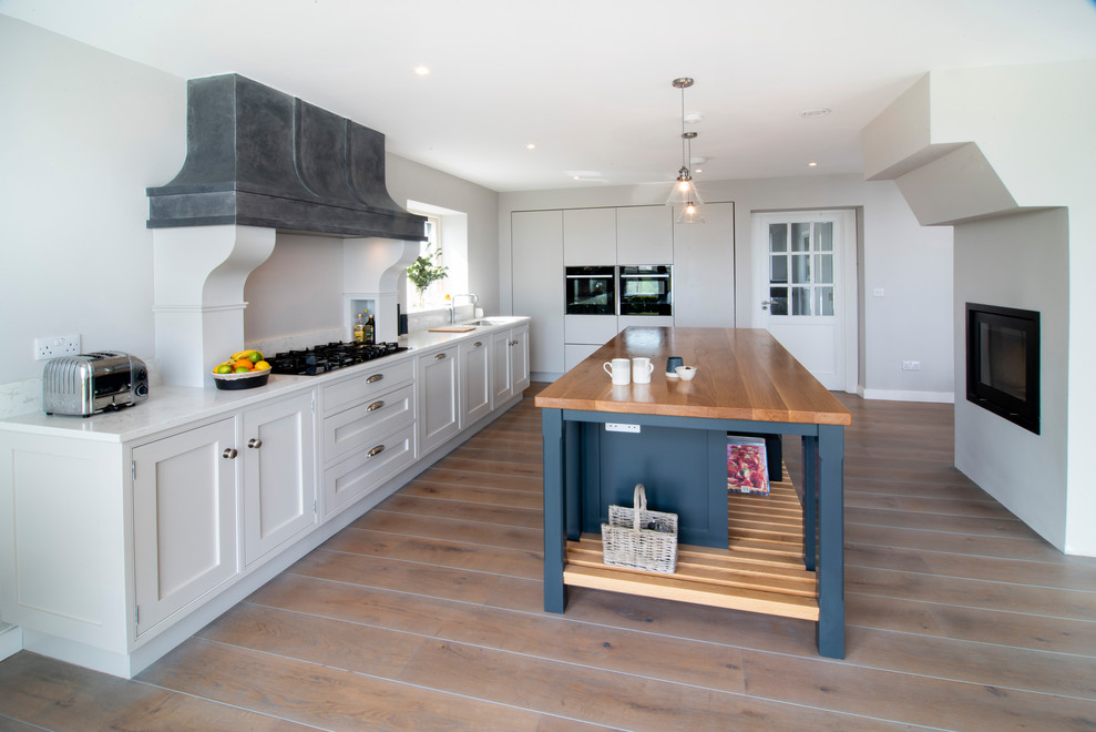 Mid-sized eclectic l-shaped eat-in kitchen in Cork with an integrated sink, shaker cabinets, white cabinets, quartzite benchtops, white splashback, stone slab splashback, black appliances, medium hardwood floors, with island, grey floor and white benchtop.