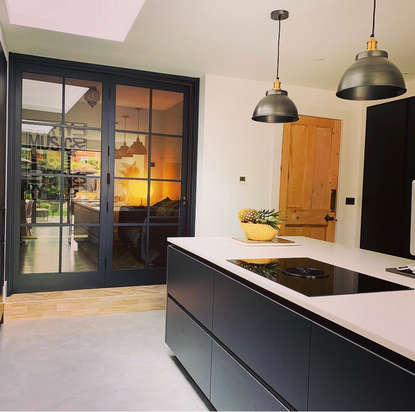 Photo of a mid-sized contemporary u-shaped open plan kitchen in Berkshire with a drop-in sink, flat-panel cabinets, black cabinets, tile benchtops, grey splashback, ceramic splashback, black appliances, concrete floors, with island, grey floor, grey benchtop and coffered.