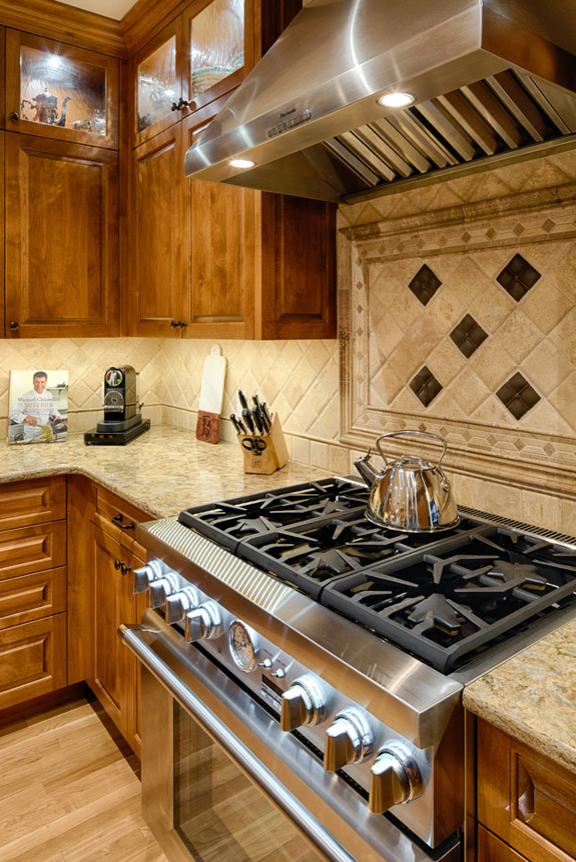 This is an example of a large traditional l-shaped eat-in kitchen in Minneapolis with an undermount sink, raised-panel cabinets, medium wood cabinets, granite benchtops, beige splashback, stone tile splashback, stainless steel appliances, light hardwood floors, with island, brown floor and yellow benchtop.