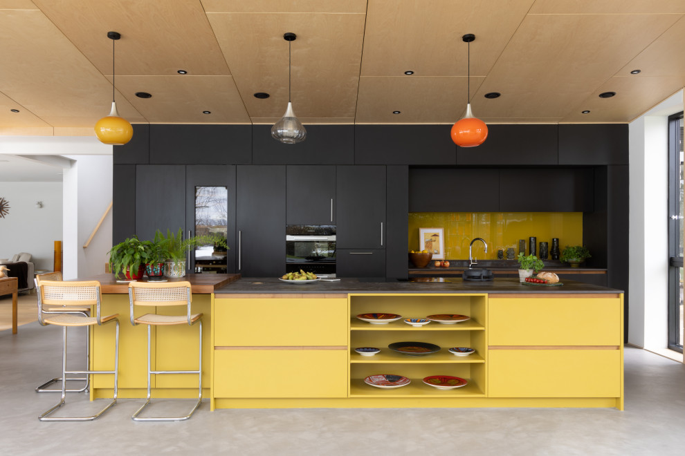 ハンプシャーにある高級な広いミッドセンチュリースタイルのおしゃれなキッチン (ドロップインシンク、フラットパネル扉のキャビネット、黄色いキャビネット、クオーツストーンカウンター、黄色いキッチンパネル、ガラスタイルのキッチンパネル、黒い調理設備、黒いキッチンカウンター、塗装板張りの天井) の写真
