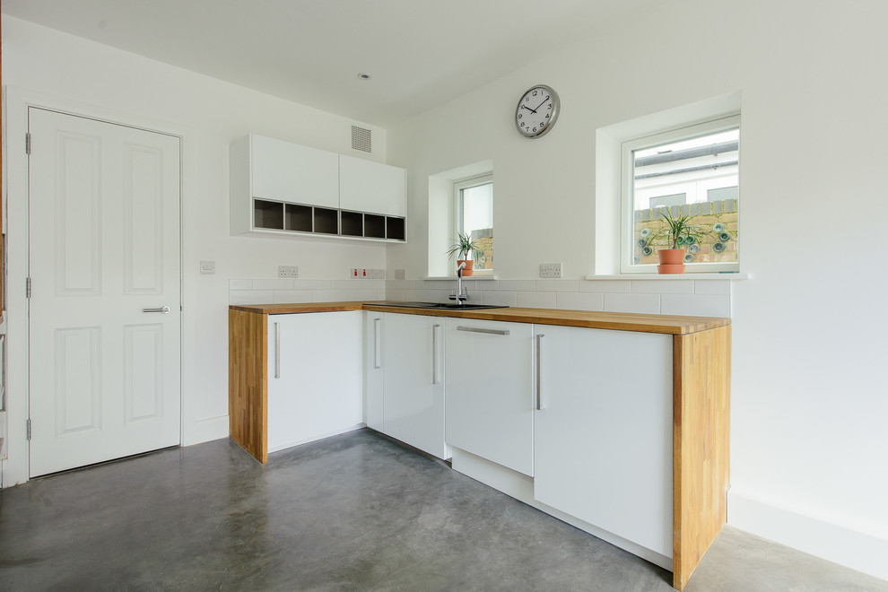 Design ideas for a mid-sized contemporary u-shaped eat-in kitchen in London with a drop-in sink, flat-panel cabinets, wood benchtops, white splashback, ceramic splashback, panelled appliances, concrete floors, no island and white cabinets.