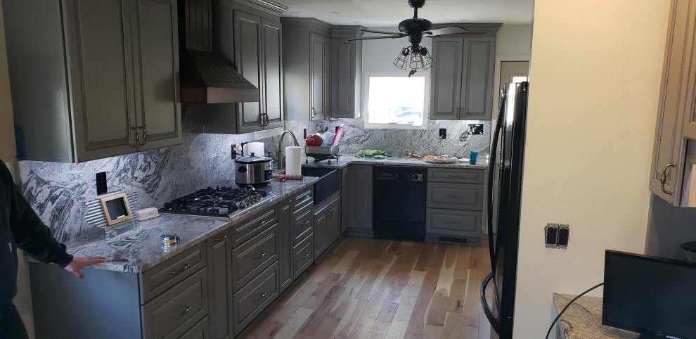 Mid-sized country galley eat-in kitchen in Bridgeport with a farmhouse sink, raised-panel cabinets, distressed cabinets, granite benchtops, grey splashback, granite splashback, black appliances, light hardwood floors, no island and grey benchtop.