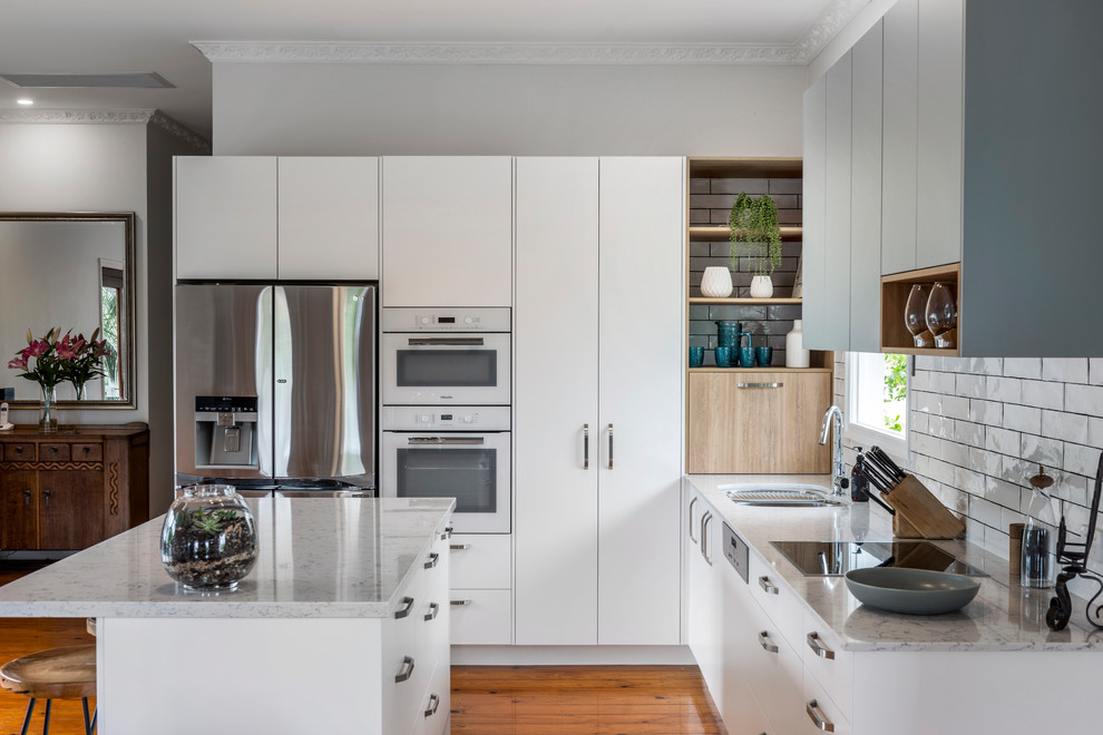 Photo of a mid-sized contemporary l-shaped open plan kitchen in Brisbane with an undermount sink, flat-panel cabinets, white cabinets, quartz benchtops, white splashback, ceramic splashback, white appliances, medium hardwood floors, with island and brown floor.