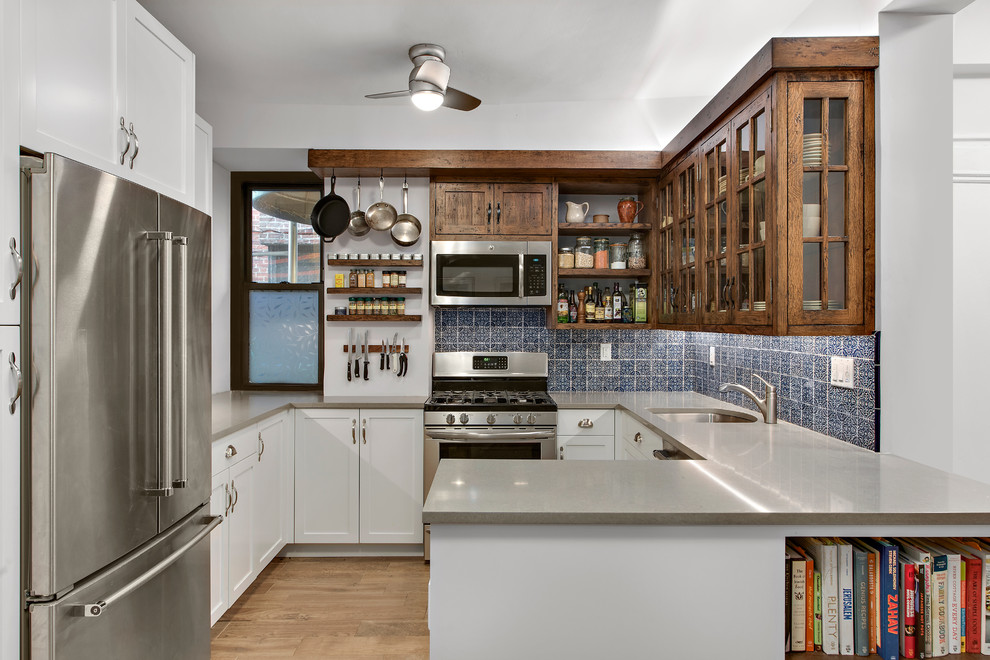 This is an example of a transitional u-shaped kitchen in New York with an undermount sink, glass-front cabinets, medium wood cabinets, blue splashback, stainless steel appliances, light hardwood floors, a peninsula and brown floor.