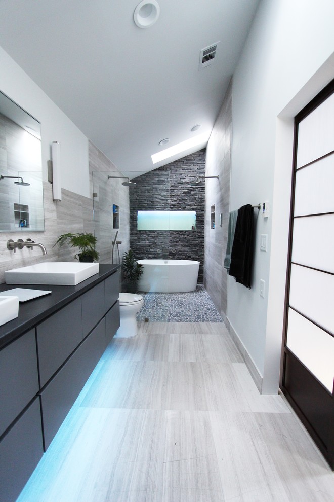 This is an example of a mid-sized contemporary master bathroom in Atlanta with flat-panel cabinets, grey cabinets, a freestanding tub, a curbless shower, gray tile, stone tile, limestone floors, white walls, a two-piece toilet, a vessel sink, granite benchtops, grey floor, an open shower, black benchtops, a niche, a double vanity, a floating vanity and vaulted.