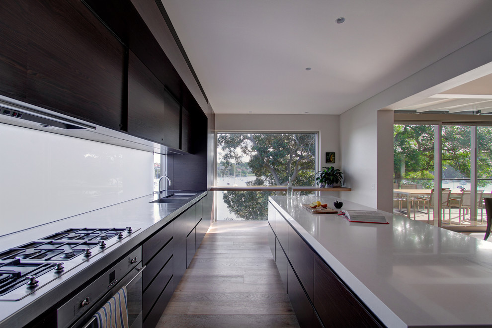 Photo of a large modern galley kitchen in Sydney with an undermount sink, flat-panel cabinets, dark wood cabinets, quartz benchtops, glass sheet splashback, stainless steel appliances, medium hardwood floors and with island.