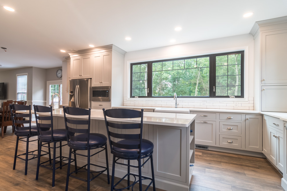 Photo of a mid-sized country l-shaped eat-in kitchen in Philadelphia with a farmhouse sink, shaker cabinets, grey cabinets, quartz benchtops, white splashback, subway tile splashback, stainless steel appliances, medium hardwood floors, with island, brown floor and grey benchtop.