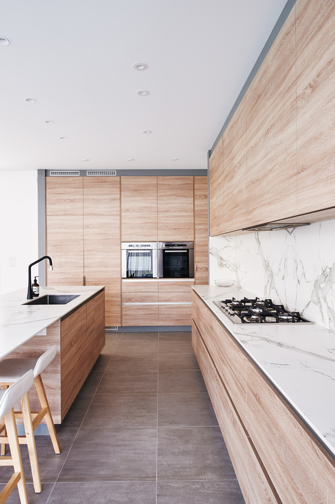 Mid-sized contemporary galley open plan kitchen in Melbourne with a drop-in sink, flat-panel cabinets, light wood cabinets, quartz benchtops, white splashback, ceramic splashback, black appliances, ceramic floors, with island, grey floor and white benchtop.