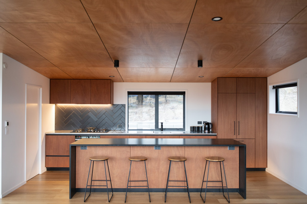 This is an example of a contemporary galley kitchen in Dunedin with flat-panel cabinets, medium wood cabinets, black splashback, panelled appliances, medium hardwood floors, with island, brown floor and black benchtop.