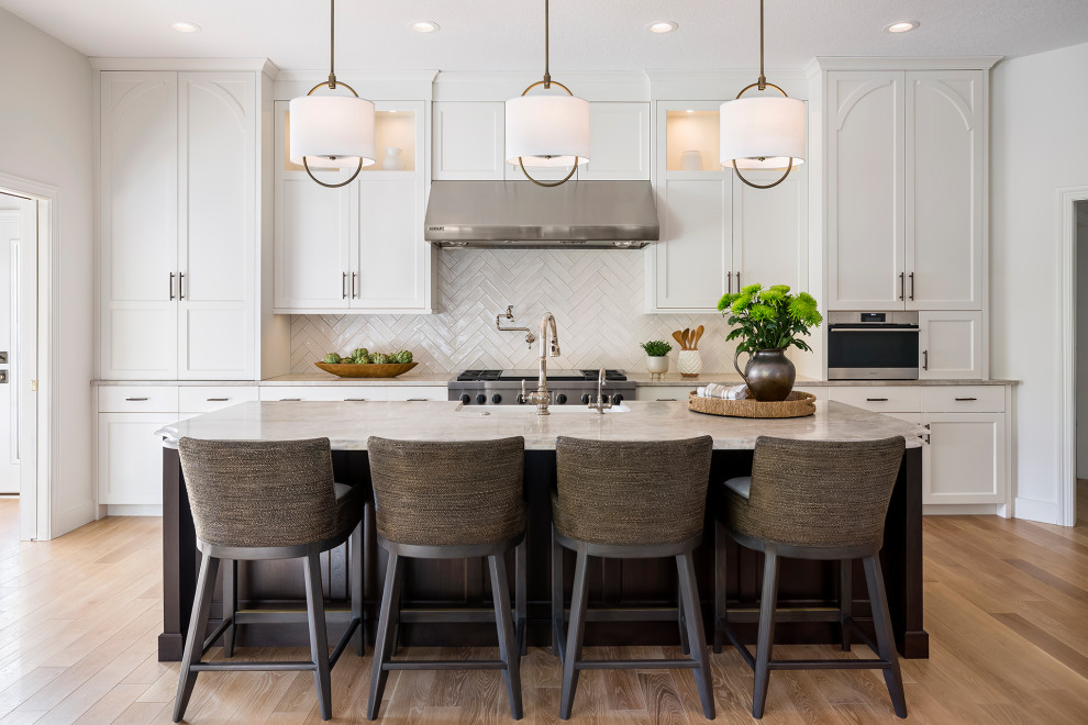 Open concept kitchen - large transitional single-wall light wood floor and brown floor open concept kitchen idea in Minneapolis with a farmhouse sink, recessed-panel cabinets, white cabinets, quartzite countertops, white backsplash, ceramic backsplash, stainless steel appliances, an island and beige countertops