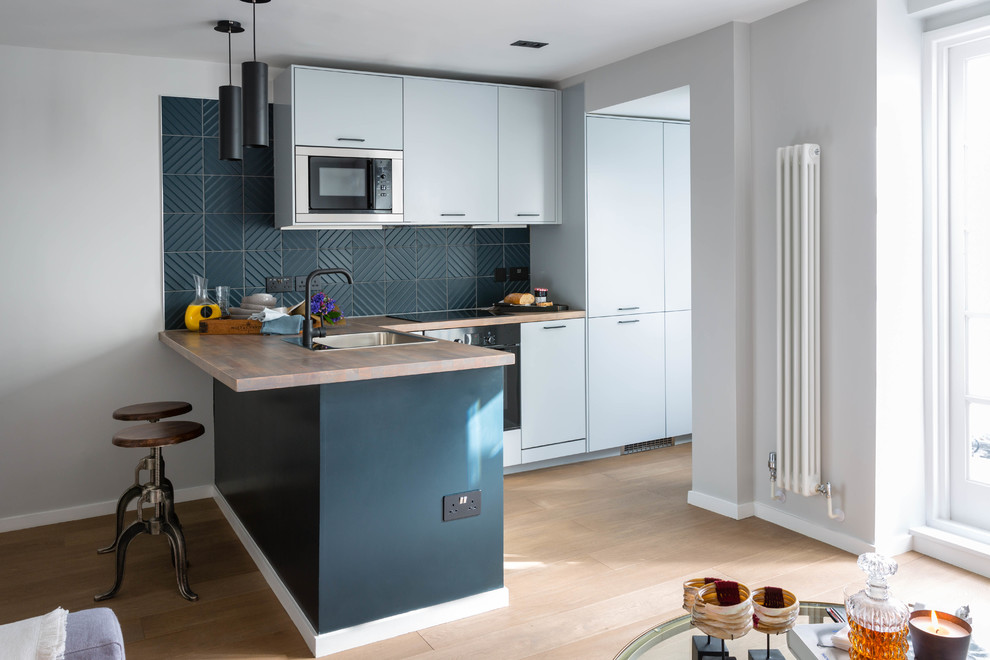 This is an example of a small contemporary l-shaped open plan kitchen in London with an undermount sink, flat-panel cabinets, wood benchtops, grey splashback, stainless steel appliances, light hardwood floors, a peninsula, beige floor and beige benchtop.