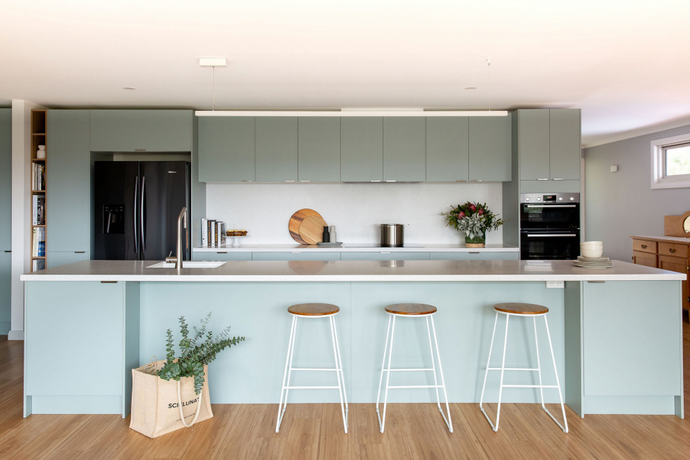 Photo of an expansive contemporary galley kitchen in Melbourne with green cabinets, quartz benchtops, white splashback, black appliances, with island, white benchtop, an undermount sink, flat-panel cabinets, medium hardwood floors and brown floor.