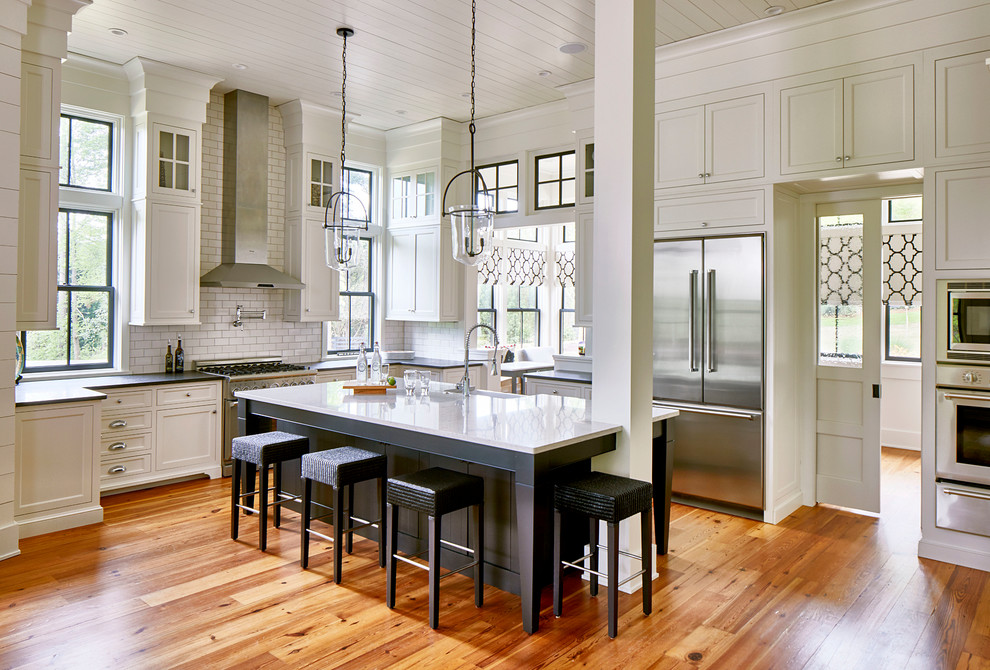 This is an example of a large country l-shaped kitchen in Atlanta with a farmhouse sink, shaker cabinets, white cabinets, white splashback, subway tile splashback, stainless steel appliances, medium hardwood floors, with island, brown floor, black benchtop and solid surface benchtops.