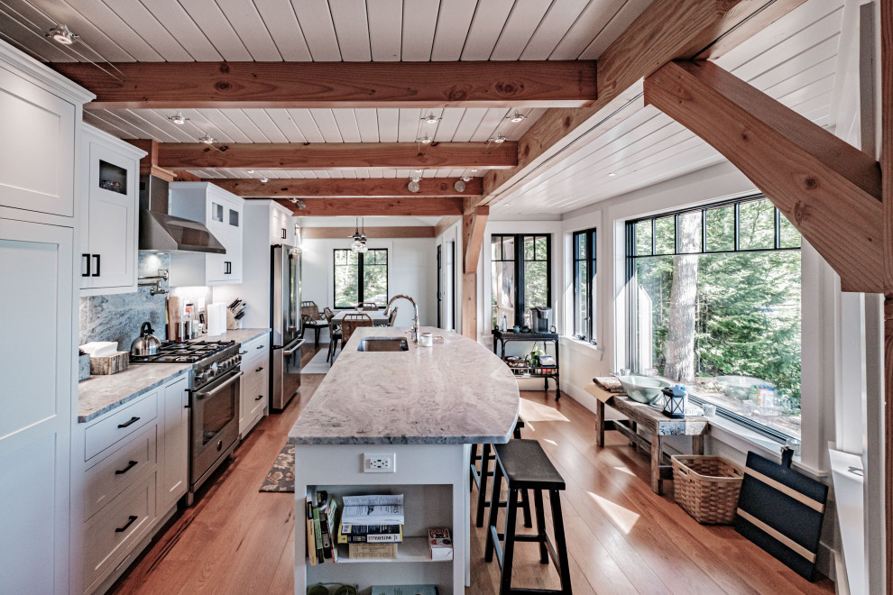 This is an example of a country galley kitchen in Portland Maine with an undermount sink, shaker cabinets, white cabinets, grey splashback, stone slab splashback, stainless steel appliances, medium hardwood floors, with island, brown floor, grey benchtop, exposed beam and timber.