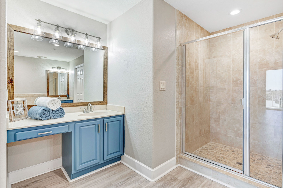 Bild på ett mellanstort maritimt beige beige en-suite badrum, med luckor med upphöjd panel, blå skåp, ett hörnbadkar, en dusch i en alkov, en toalettstol med separat cisternkåpa, beige kakel, keramikplattor, grå väggar, laminatgolv, ett integrerad handfat, marmorbänkskiva, grått golv och dusch med gångjärnsdörr