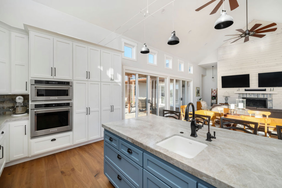 Photo of a large country u-shaped open plan kitchen in San Luis Obispo with a drop-in sink, recessed-panel cabinets, white cabinets, marble benchtops, multi-coloured splashback, granite splashback, stainless steel appliances, light hardwood floors, with island, brown floor, grey benchtop and vaulted.