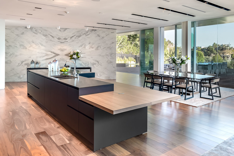 Design ideas for an expansive modern galley open plan kitchen in Los Angeles with an undermount sink, flat-panel cabinets, light wood cabinets, stainless steel benchtops, grey splashback, stainless steel appliances, light hardwood floors and with island.