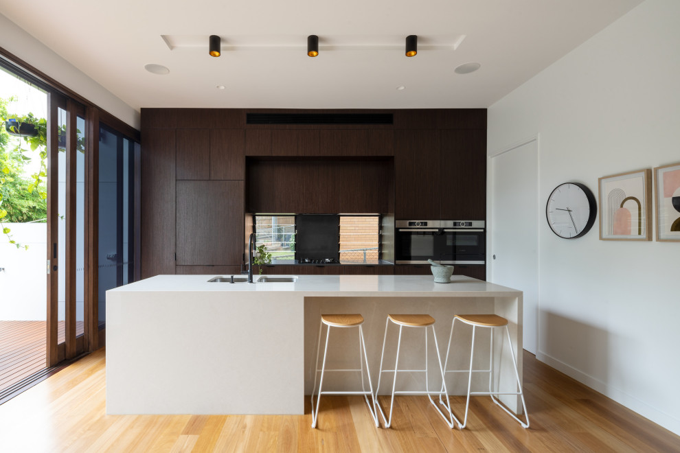 This is an example of a contemporary galley open plan kitchen in Brisbane with a double-bowl sink, flat-panel cabinets, dark wood cabinets, quartz benchtops, window splashback, panelled appliances, medium hardwood floors, with island, brown floor and white benchtop.