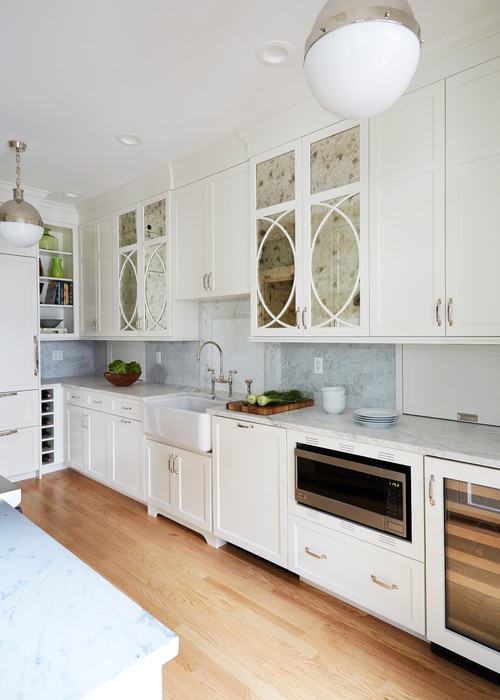Bright white kitchen with shaker-style RTA cabinets, mirrored glass uppers with oval mullions, apron-front sink, marble countertops, and built-in microwave/wine cooler on light wood floors.