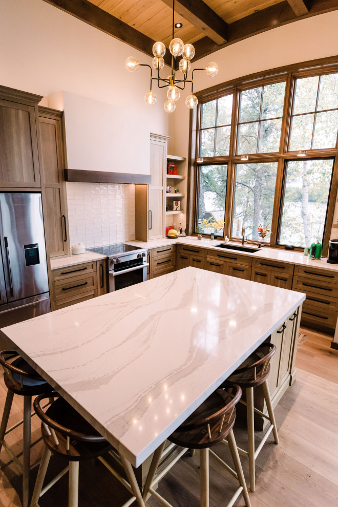 Example of a large l-shaped medium tone wood floor, brown floor and exposed beam open concept kitchen design in Other with shaker cabinets, brown cabinets, quartzite countertops, white backsplash, subway tile backsplash, stainless steel appliances, an island and white countertops