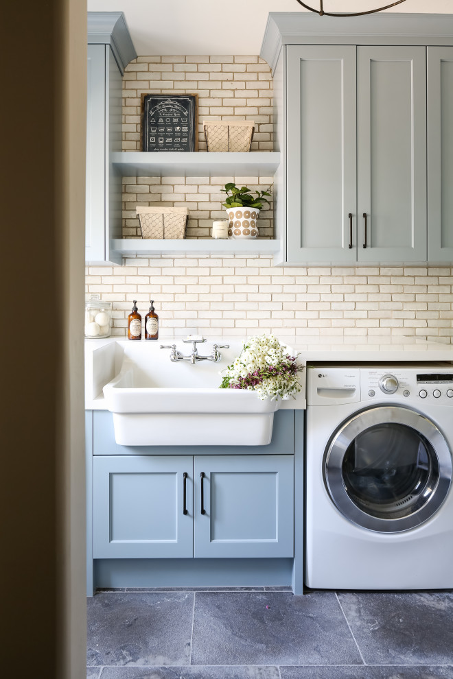 This is an example of a mid-sized transitional single-wall dedicated laundry room in San Diego with quartz benchtops, slate floors, white benchtop, a farmhouse sink, shaker cabinets, grey cabinets, a side-by-side washer and dryer and grey floor.