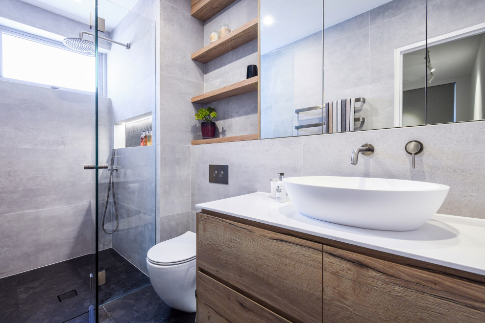 Exempel på ett mellanstort modernt vit vitt badrum med dusch, med släta luckor, skåp i mellenmörkt trä, en öppen dusch, en vägghängd toalettstol, svart kakel, grå kakel, keramikplattor, grå väggar, klinkergolv i keramik, ett nedsänkt handfat, bänkskiva i akrylsten, svart golv och dusch med gångjärnsdörr