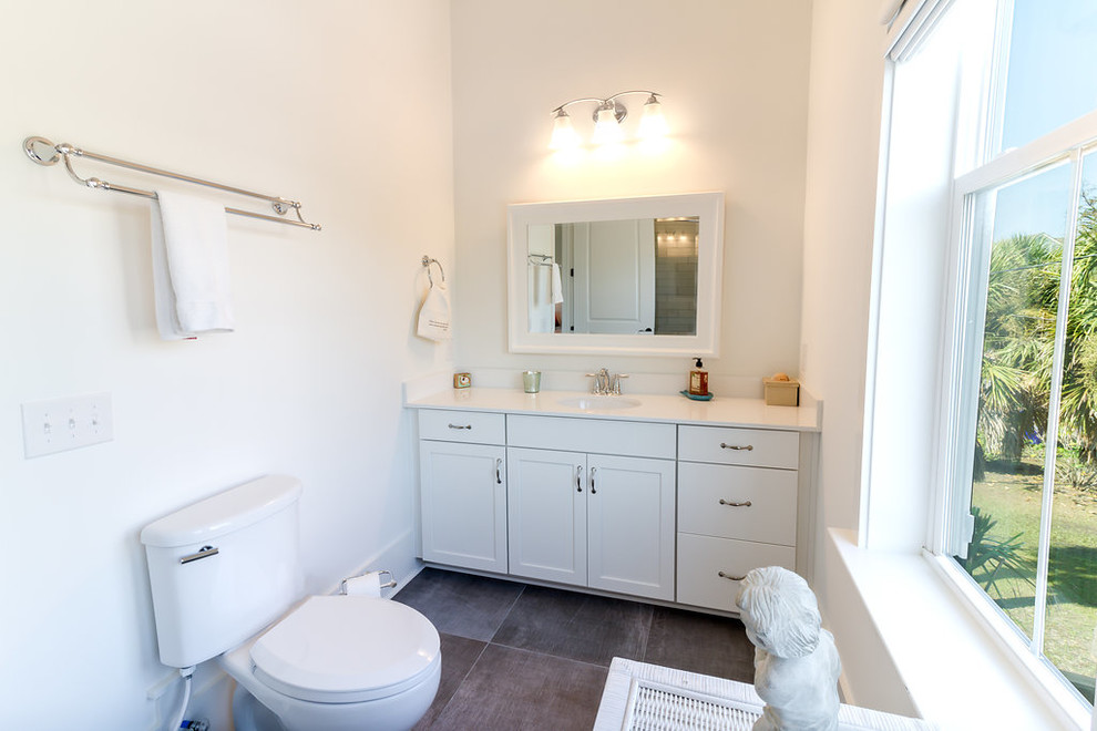 Photo of a mid-sized beach style 3/4 bathroom in Charleston with shaker cabinets, white cabinets, an alcove shower, a two-piece toilet, white tile, subway tile, white walls, porcelain floors, an undermount sink, solid surface benchtops, grey floor, a hinged shower door and white benchtops.