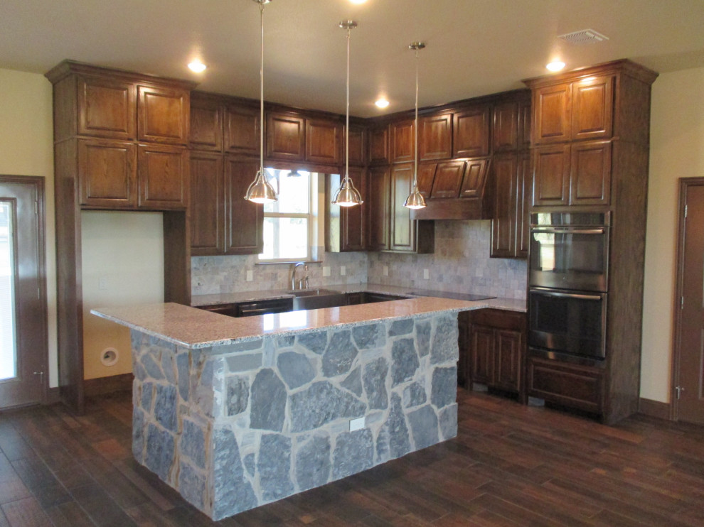 This is an example of a mid-sized arts and crafts l-shaped kitchen in Dallas with a farmhouse sink, raised-panel cabinets, dark wood cabinets, grey splashback, stone tile splashback, black appliances, dark hardwood floors, with island, brown floor and grey benchtop.