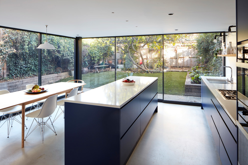 Photo of a large contemporary galley open plan kitchen in London with a drop-in sink, flat-panel cabinets, blue cabinets, quartzite benchtops, white splashback, ceramic splashback, stainless steel appliances, concrete floors, with island, grey floor and white benchtop.