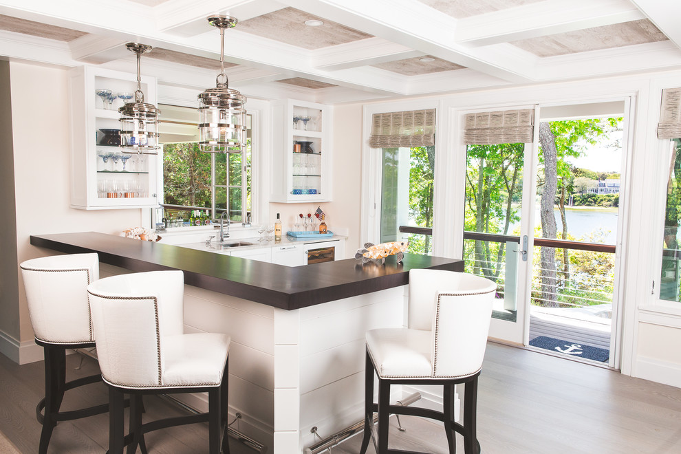 Photo of a beach style seated home bar in Boston with an undermount sink, glass-front cabinets, white cabinets, light hardwood floors, beige floor and brown benchtop.