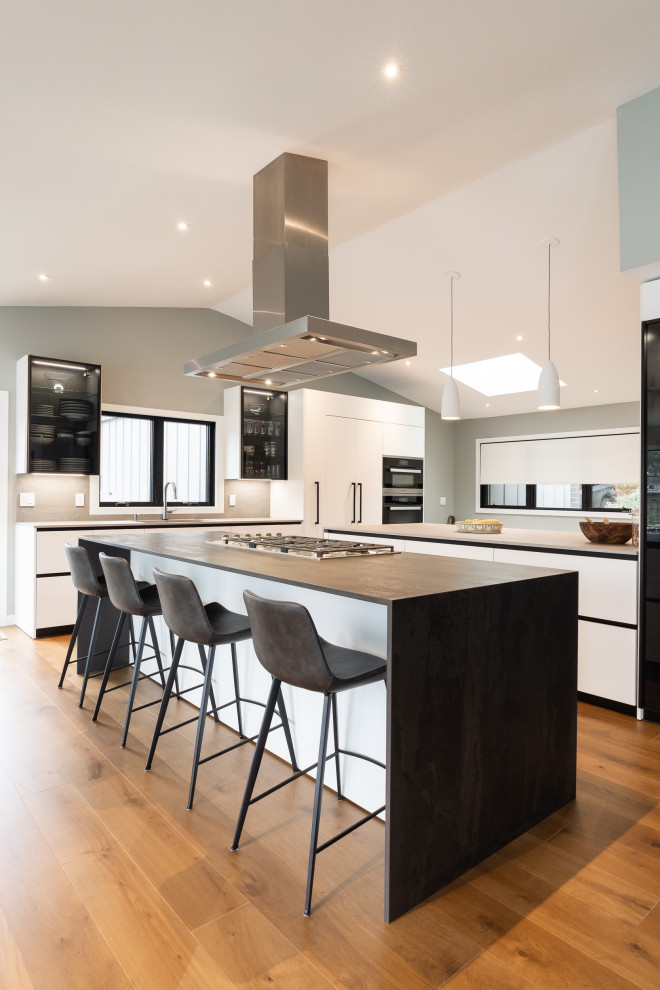 Photo of an expansive contemporary single-wall open plan kitchen in Portland with an undermount sink, flat-panel cabinets, white cabinets, quartz benchtops, grey splashback, engineered quartz splashback, panelled appliances, medium hardwood floors, multiple islands, brown floor, grey benchtop and vaulted.