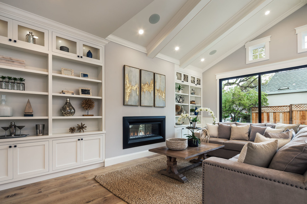 Inspiration for a mid-sized country open concept family room in San Francisco with a library, grey walls, light hardwood floors, a two-sided fireplace, a plaster fireplace surround, a wall-mounted tv and grey floor.