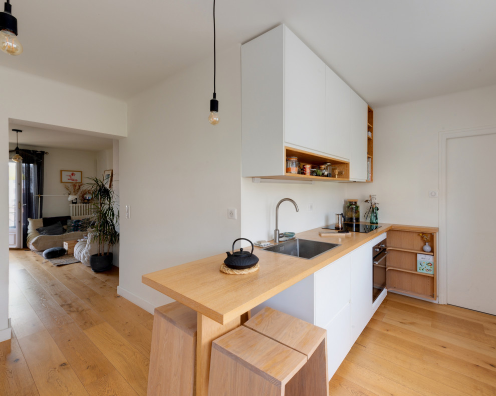 Photo of a mid-sized scandinavian single-wall open plan kitchen in Other with a single-bowl sink, beaded inset cabinets, white cabinets, wood benchtops, white splashback, black appliances, light hardwood floors, a peninsula, brown floor and brown benchtop.