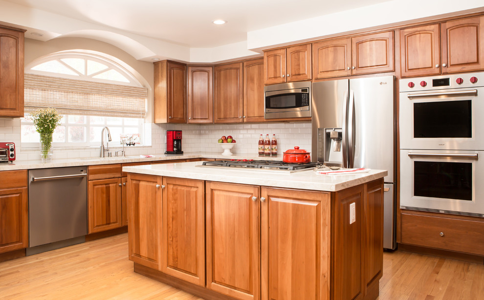 Mid-sized traditional l-shaped open plan kitchen in Los Angeles with an undermount sink, raised-panel cabinets, medium wood cabinets, marble benchtops, white splashback, subway tile splashback, stainless steel appliances, light hardwood floors and with island.
