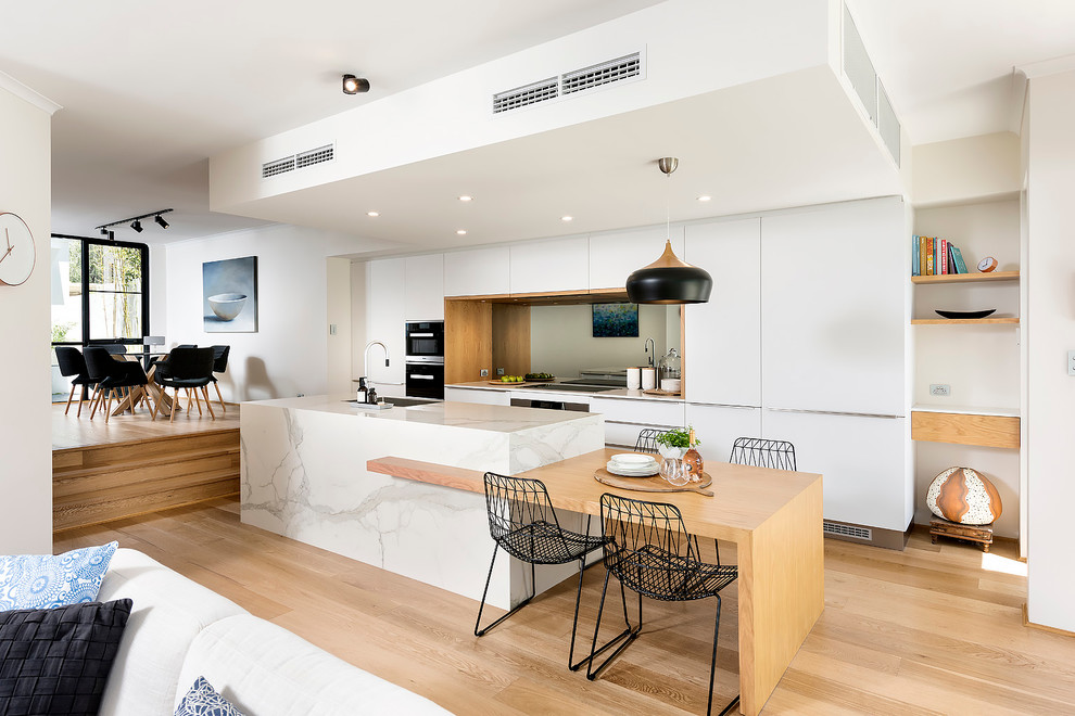 Photo of a large scandinavian single-wall open plan kitchen in Perth with flat-panel cabinets, white cabinets, mirror splashback, black appliances, light hardwood floors, with island, a double-bowl sink, tile benchtops, metallic splashback, brown floor and white benchtop.