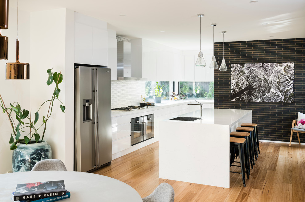 Large contemporary l-shaped eat-in kitchen in Melbourne with a drop-in sink, flat-panel cabinets, white cabinets, quartz benchtops, white splashback, cement tile splashback, black appliances, medium hardwood floors and with island.