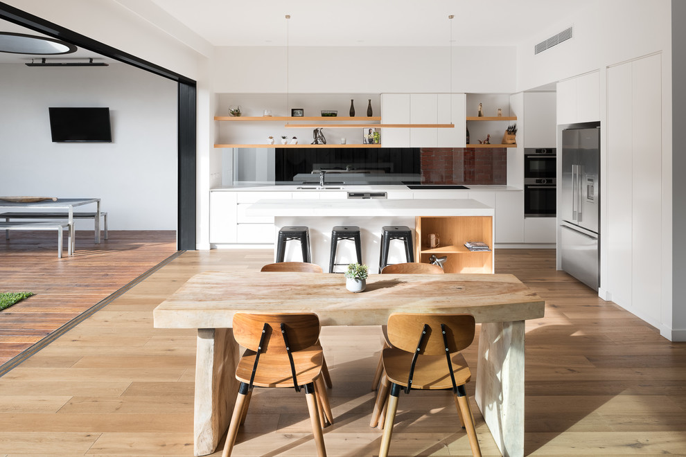 Contemporary eat-in kitchen in Melbourne with a double-bowl sink, flat-panel cabinets, white cabinets, black splashback, glass sheet splashback, stainless steel appliances, light hardwood floors, with island and beige floor.