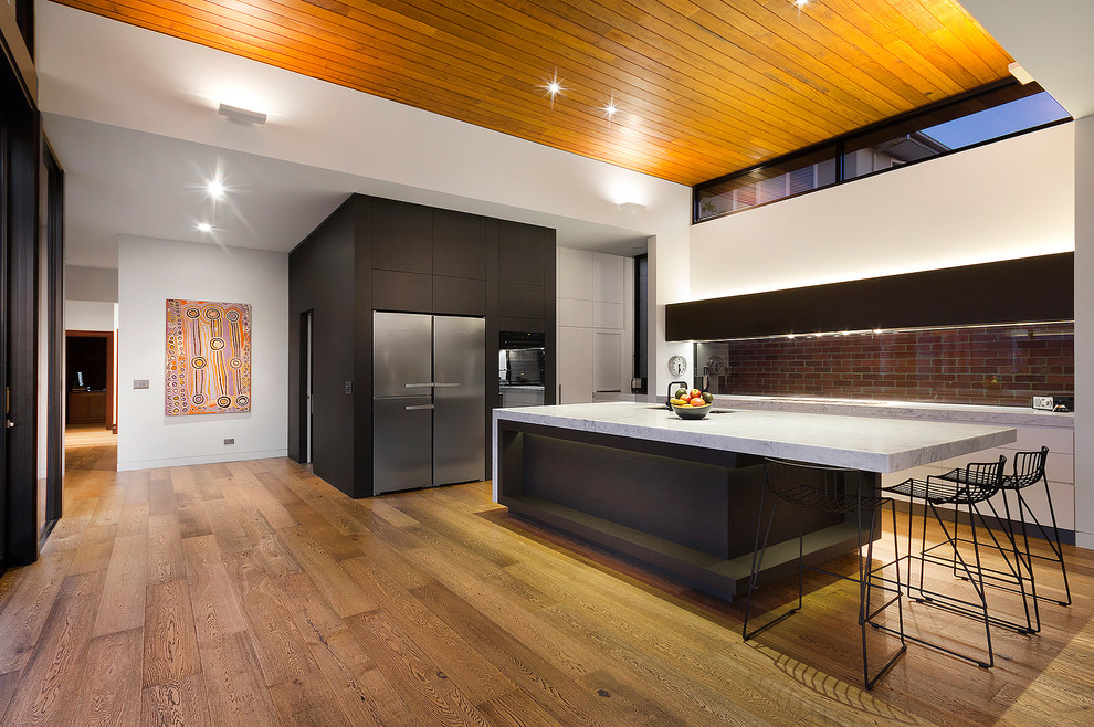 Inspiration for a large contemporary l-shaped open plan kitchen in Melbourne with flat-panel cabinets, white cabinets, marble benchtops, stainless steel appliances, medium hardwood floors, with island, brown splashback and brick splashback.