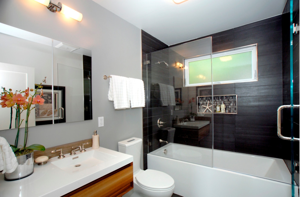 Mid-sized modern 3/4 bathroom in San Francisco with flat-panel cabinets, medium wood cabinets, an alcove tub, a shower/bathtub combo, a one-piece toilet, black tile, grey walls, an integrated sink, engineered quartz benchtops, a hinged shower door and white benchtops.