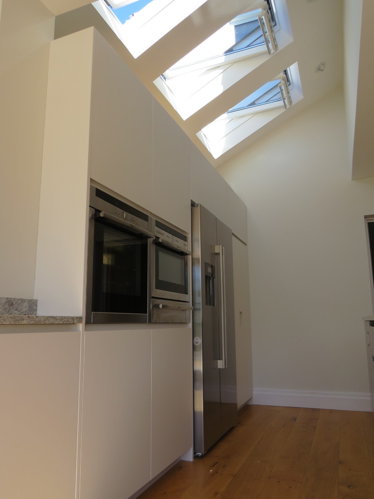 Large contemporary l-shaped home with a built-in sink, flat-panel cabinets, white cabinets, granite worktops, green splashback, stone slab splashback, stainless steel appliances, light hardwood flooring and an island.