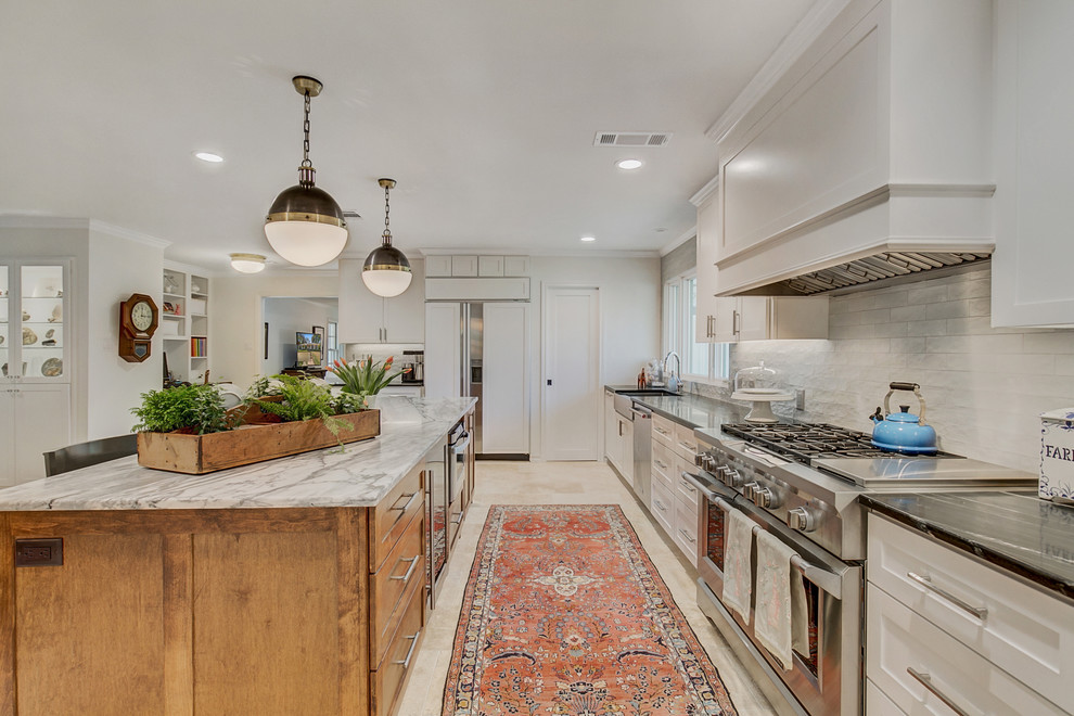 Large eclectic l-shaped open plan kitchen in Dallas with a farmhouse sink, shaker cabinets, white cabinets, soapstone benchtops, grey splashback, ceramic splashback, stainless steel appliances, travertine floors, with island, beige floor and black benchtop.