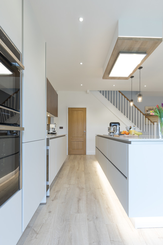 This is an example of a mid-sized modern single-wall open plan kitchen in Hampshire with an undermount sink, flat-panel cabinets, white cabinets, quartzite benchtops, metallic splashback, panelled appliances, light hardwood floors and with island.